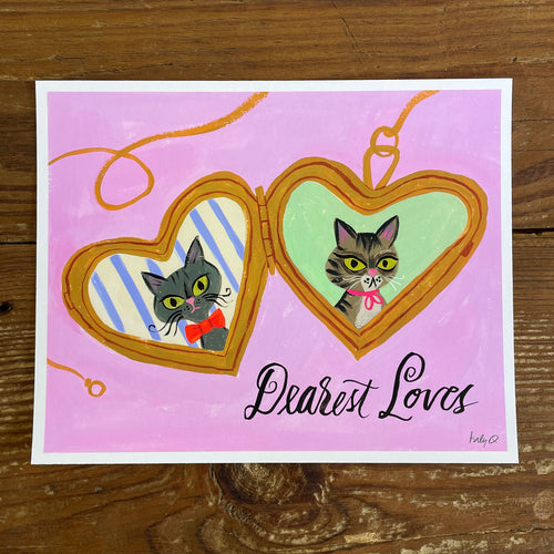 Illustration of a Russian Blue & tabby cat  inside a heart-shaped gold locket on a pink background, hand lettering reads Dearest Loves