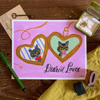 Illustration of a Russian Blue & tabby cat  inside a heart-shaped gold locket on a pink background, hand lettering reads Dearest Loves